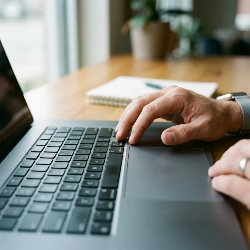 A macro editorial photo of a person resting their hand on a sleek laptop trackpad at a wooden desk